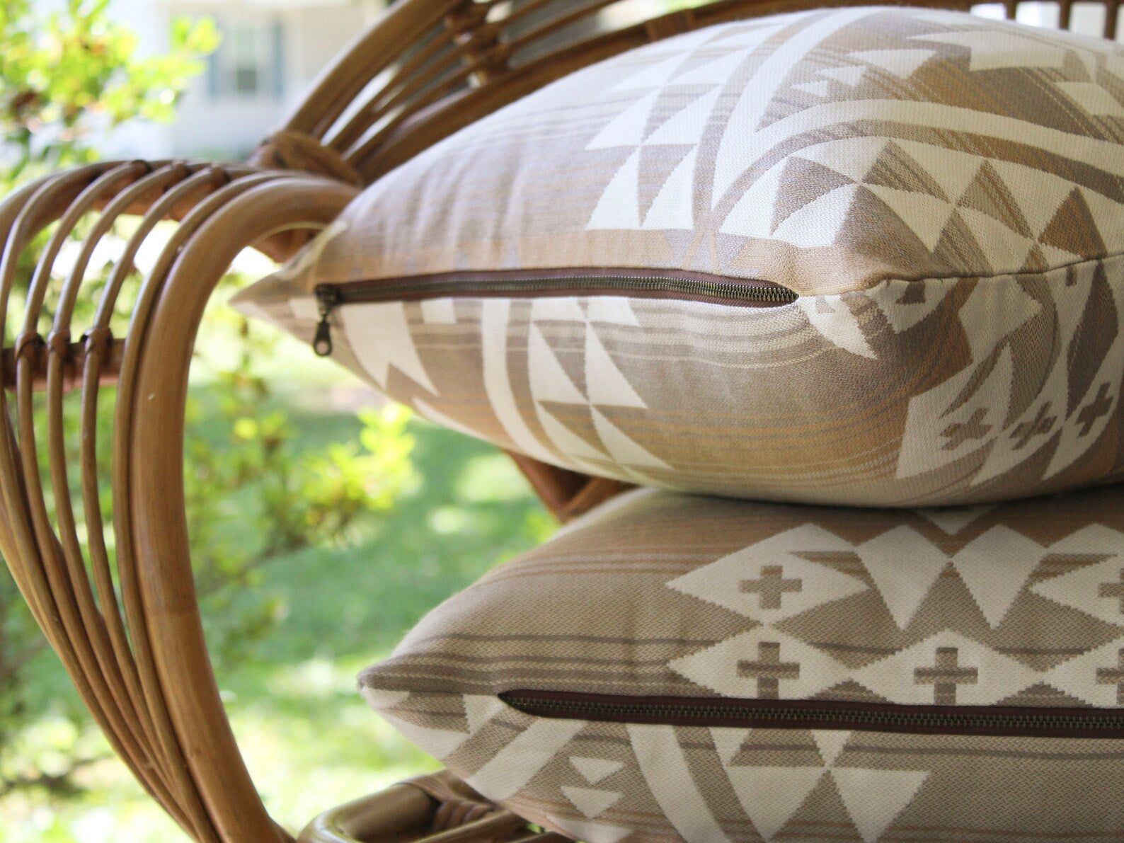 Close up of the exposed metal zipper on two outdoor pillow covers, stacked on a wicker chair with a blurred green outdoor background. The outdoor pillow covers are in the cream and tan southwestern Canyonlands pattern.