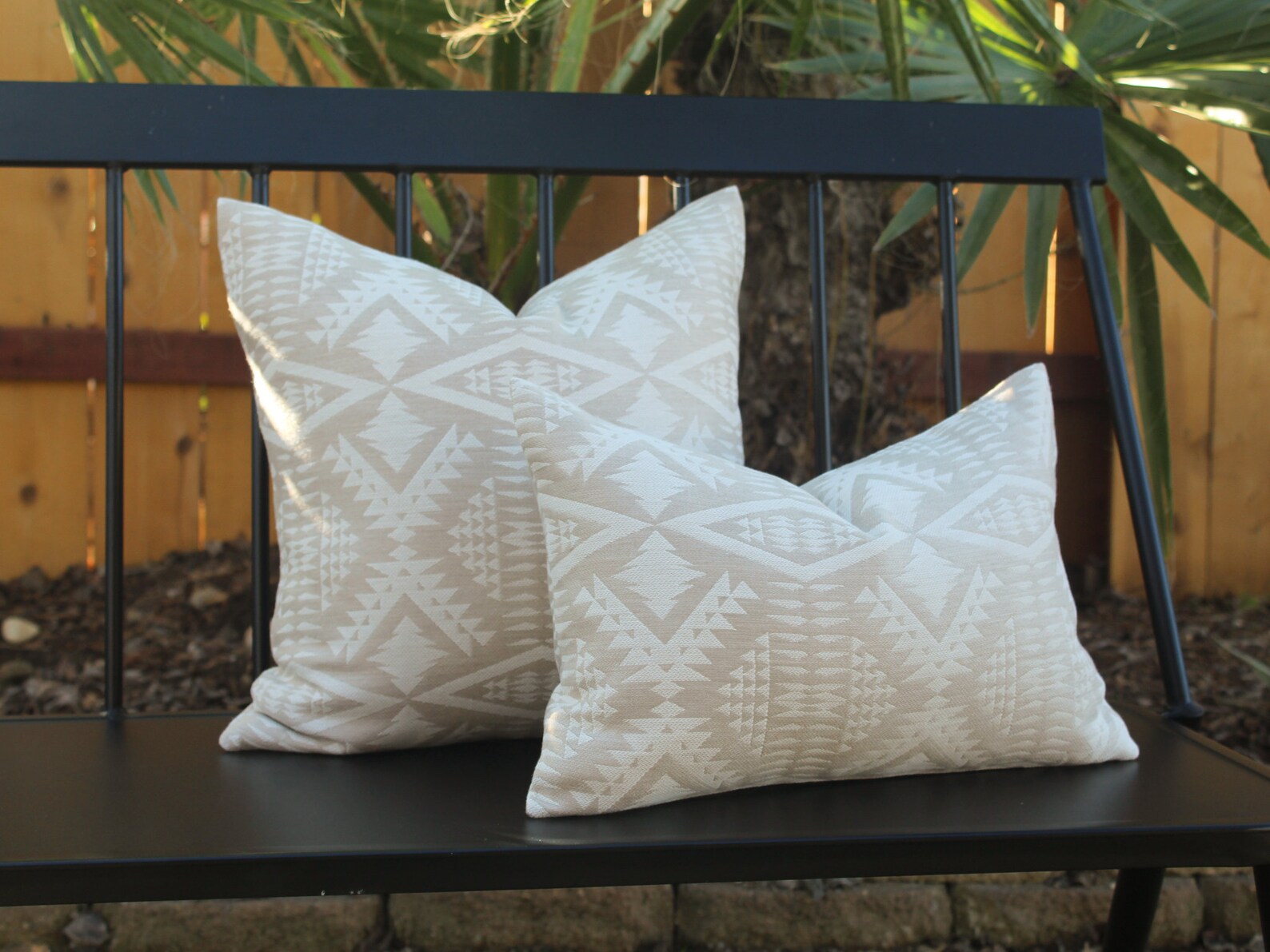 Two outdoor pillows in the two-tone tan River Tonal Sand pattern on a black metal bench with a blurred natural background