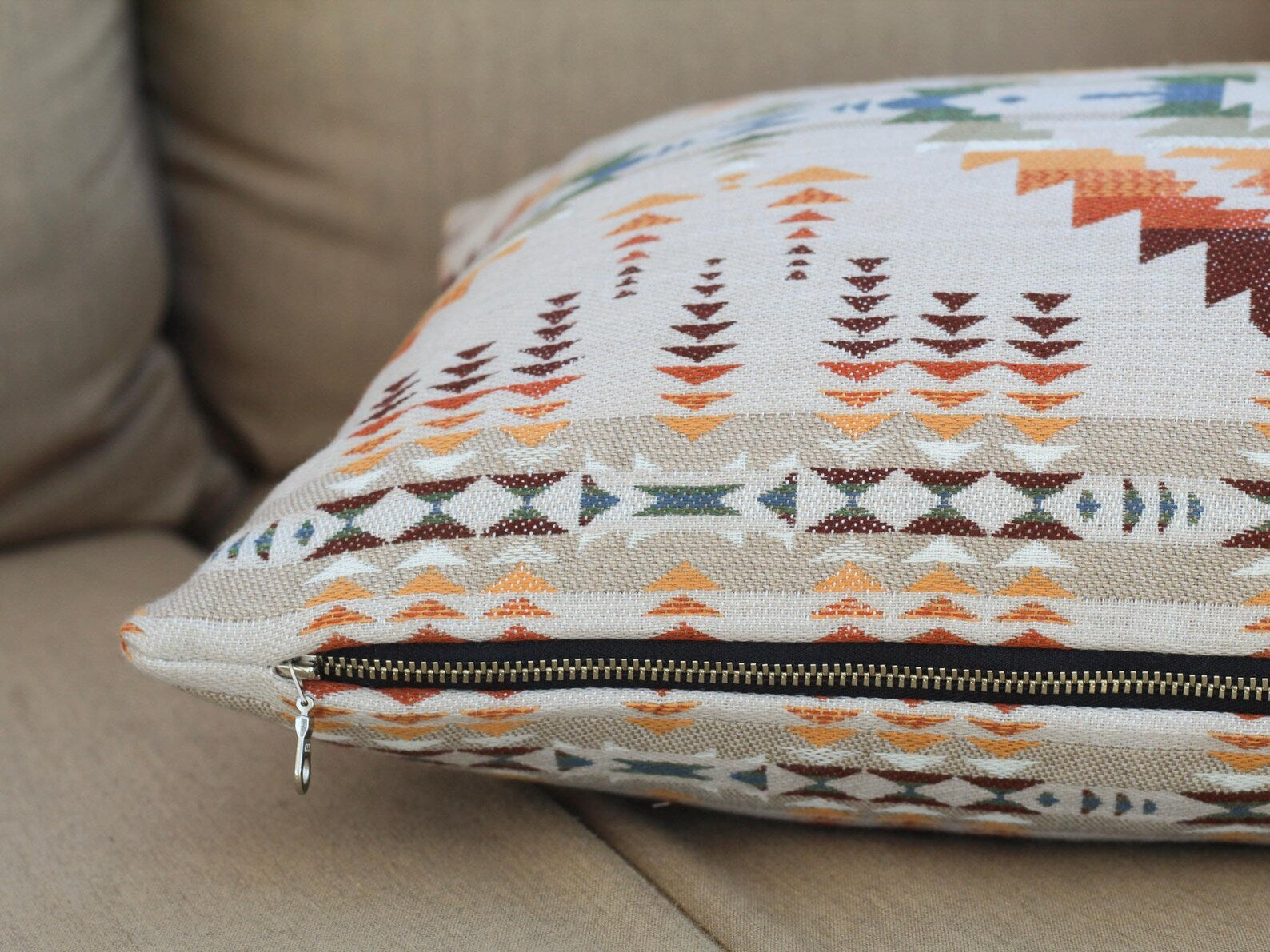 Close up of zipper on outdoor pillow covers in the Juniper Mesa Adobe pattern on a beige outdoor sofa. Juniper Mesa Adobe as a beige background with a southwestern pattern in shades of brown, red, orange, yellow, green, and blue.