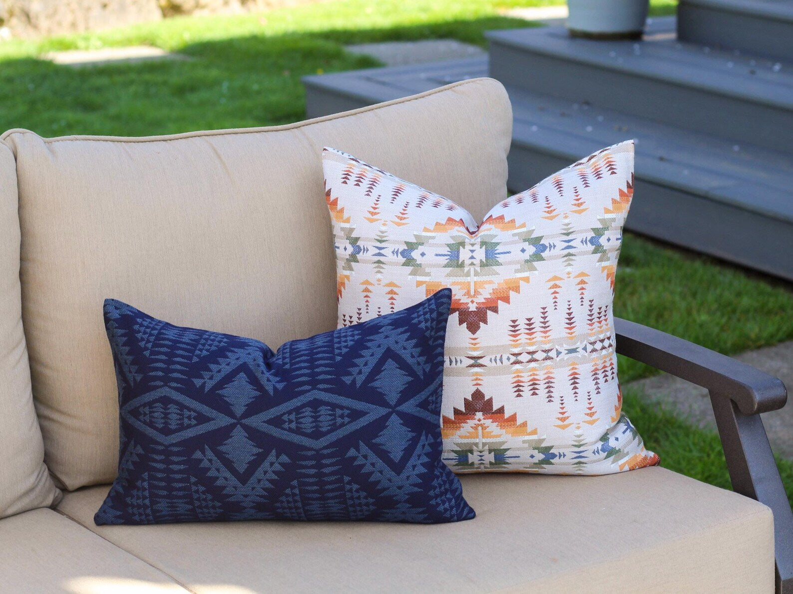 Two outdoor pillow covers in the Juniper Mesa Adobe and River Tonal Midnight patterns on a beige outdoor sofa. Juniper Mesa Adobe as a beige background with a southwestern pattern in shades of brown, red, orange, yellow, green, and blue.