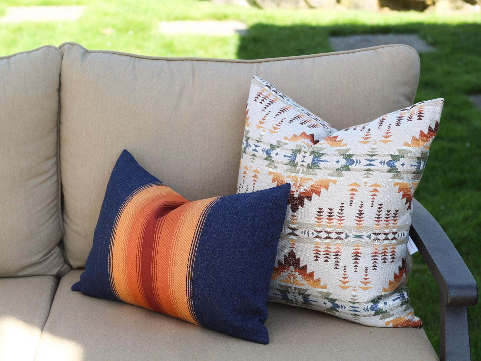 Two outdoor pillow covers in the Juniper Mesa Adobe and Grand Canyon National Park patterns on a beige outdoor sofa. Juniper Mesa Adobe as a beige background with a southwestern pattern in shades of brown, red, orange, yellow, green, and blue.