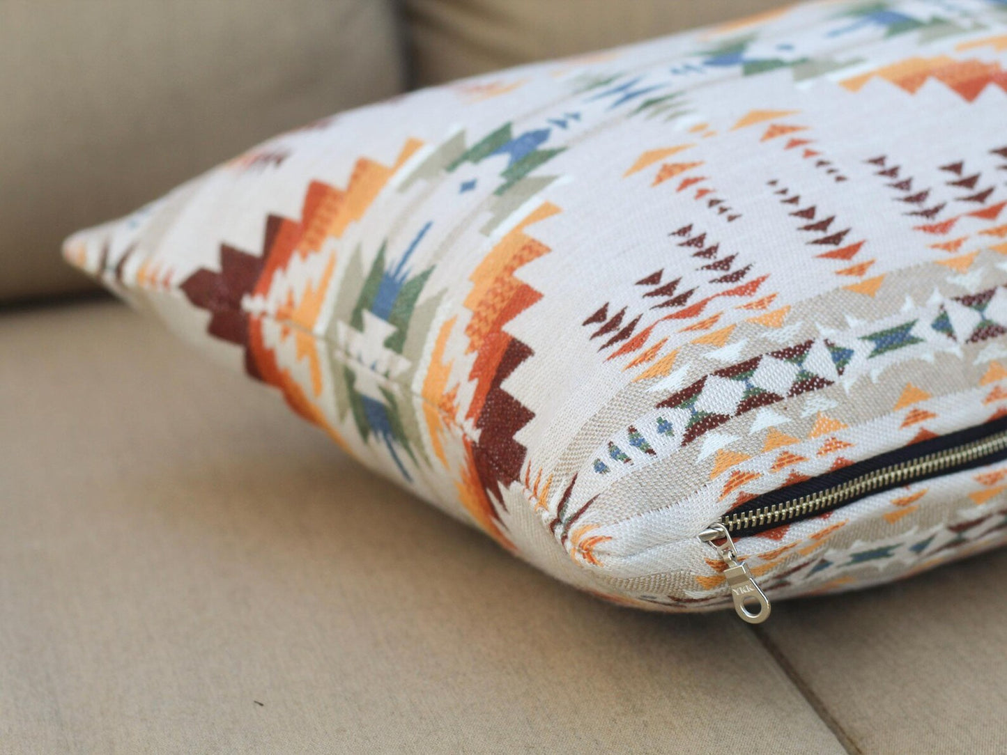 Close up of zipper on outdoor pillow covers in the Juniper Mesa Adobe pattern on a beige outdoor sofa. Juniper Mesa Adobe as a beige background with a southwestern pattern in shades of brown, red, orange, yellow, green, and blue.