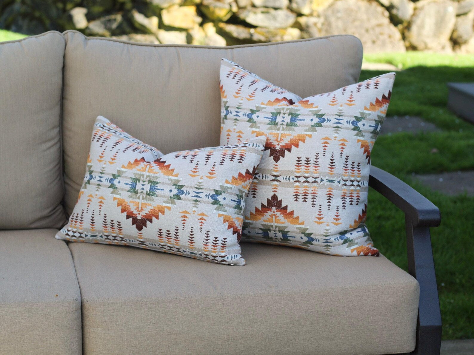 Two outdoor pillow covers in the Juniper Mesa Adobe pattern on a beige outdoor sofa. Juniper Mesa Adobe as a beige background with a southwestern pattern in shades of brown, red, orange, yellow, green, and blue.