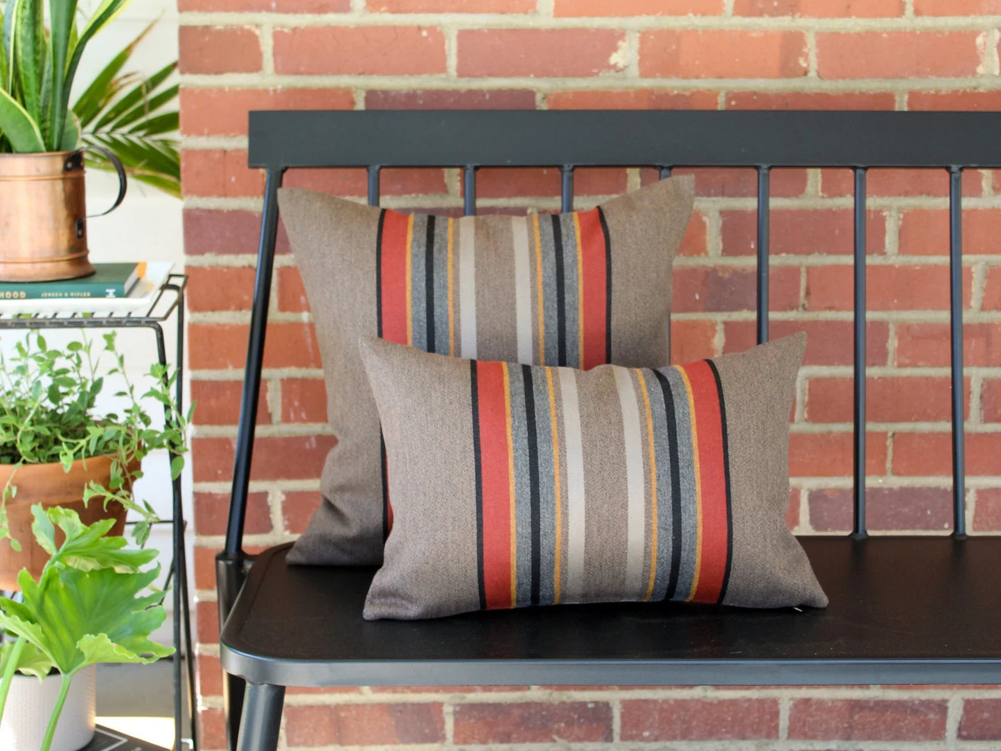 Two outdoor pillows in the Yakima Park Umber stripe on a black metal bench with a brick wall background