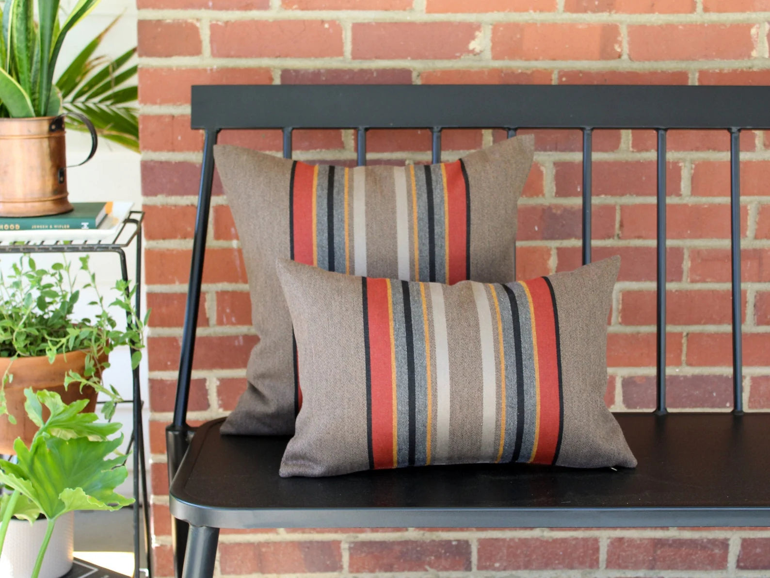 Two outdoor pillows in the Yakima Park Umber stripe on a black metal bench with a brick wall background