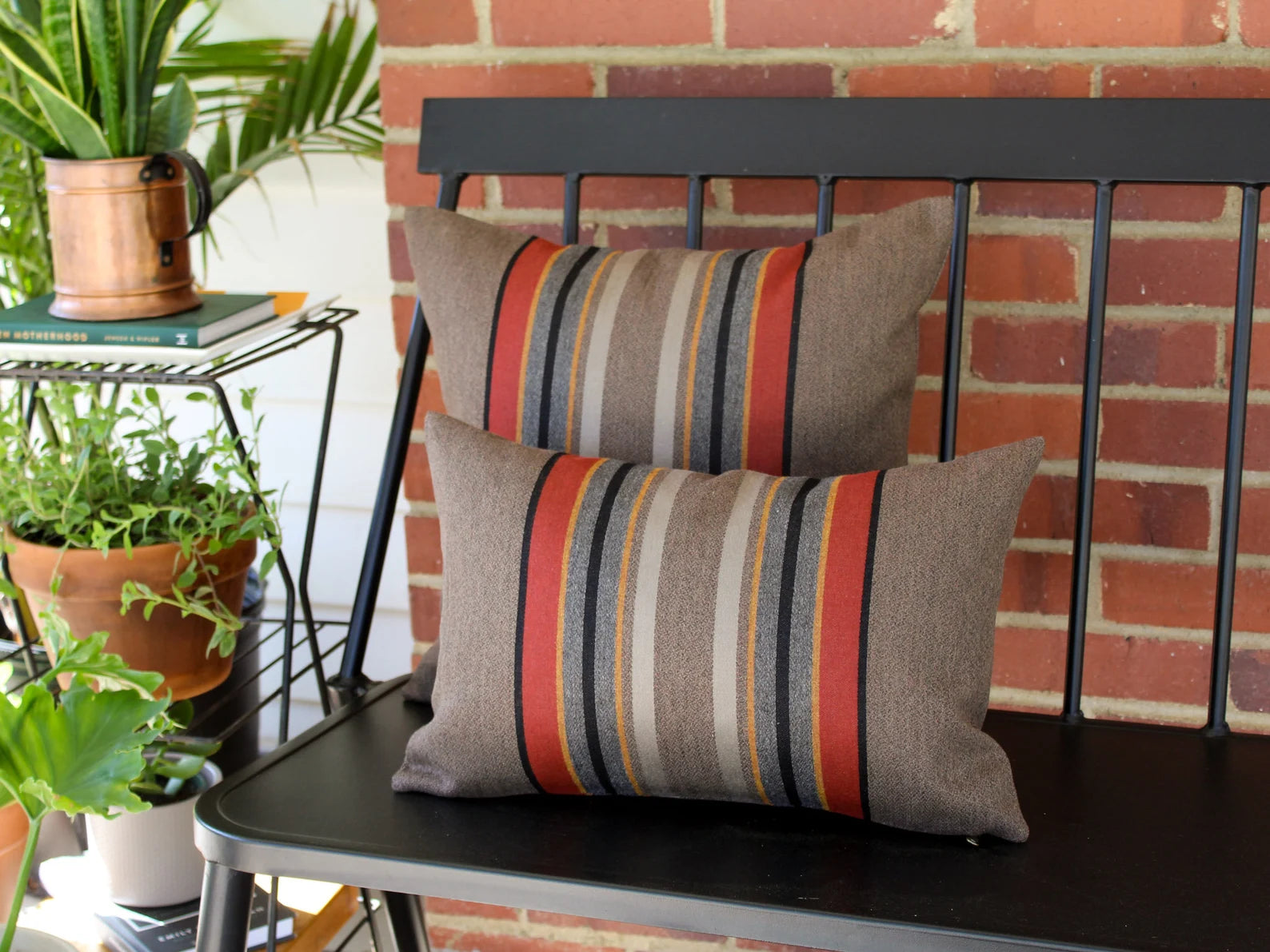 Outdoor pillows in the Yakima Park Umber stripe on a black metal bench with plants and a brick wall in the background