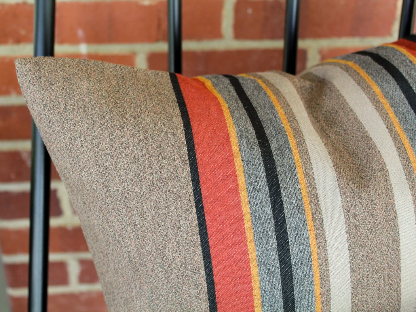 Outdoor pillow with red, black, blue, gold and gray stripes against a brick wall.
