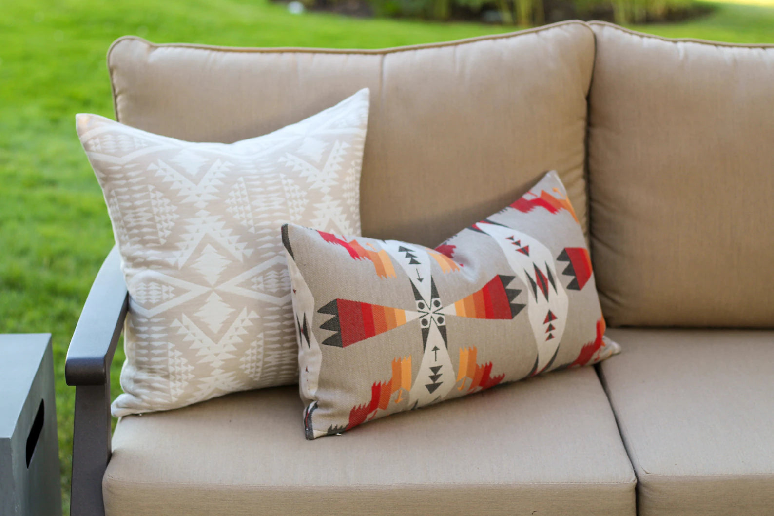 Two outdoor pillows in the two-tone tan River Tonal Sand pattern and southwestern Tucson Hawk pattern on a beige outdoor sofa with a grassy background