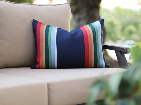 Colorful striped pillow with navy background and stripes in red, orange, yellow, green, blue, and white on a beige outdoor sofa with blurred greenery in the background