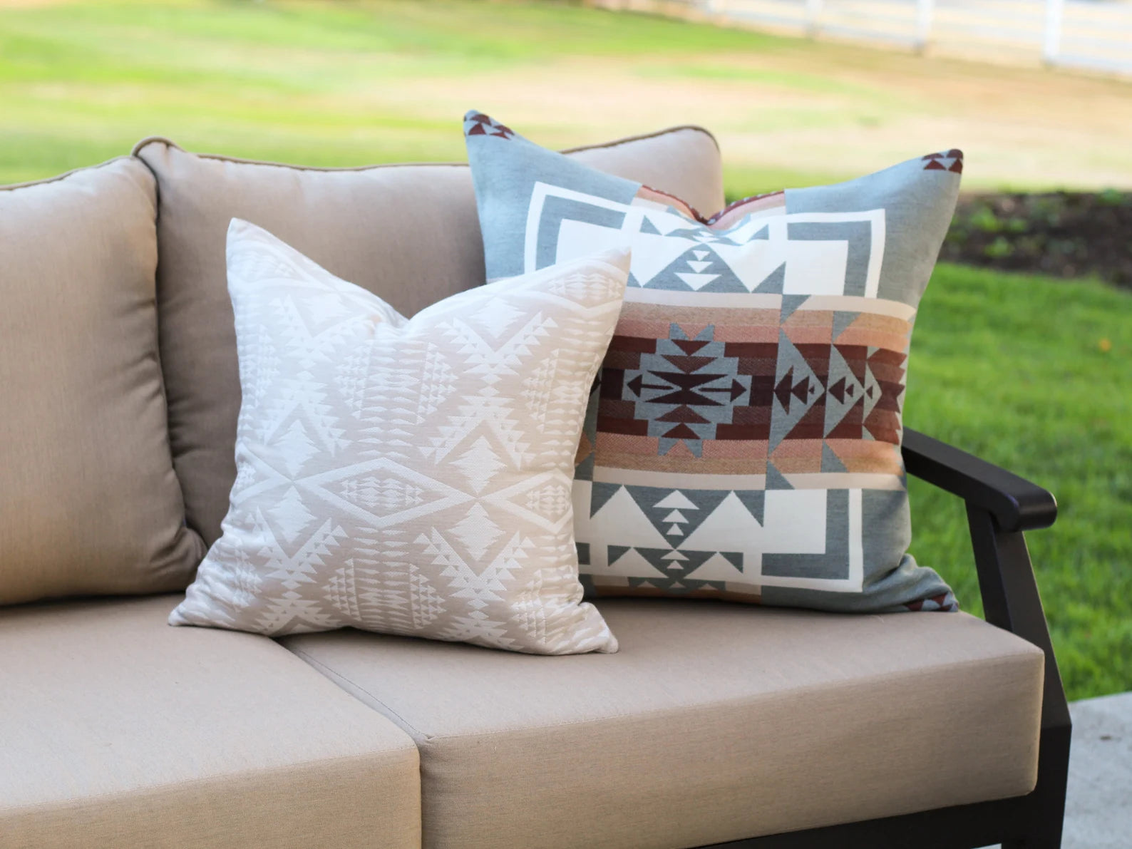 Beige outdoor sofa with outdoor pillows on a grassy background. One pillow is the two-tone tan River Tonal Sand pattern and the other is the blue, brown, and white southwestern Chief Joseph pattern on a bench with a natural background.