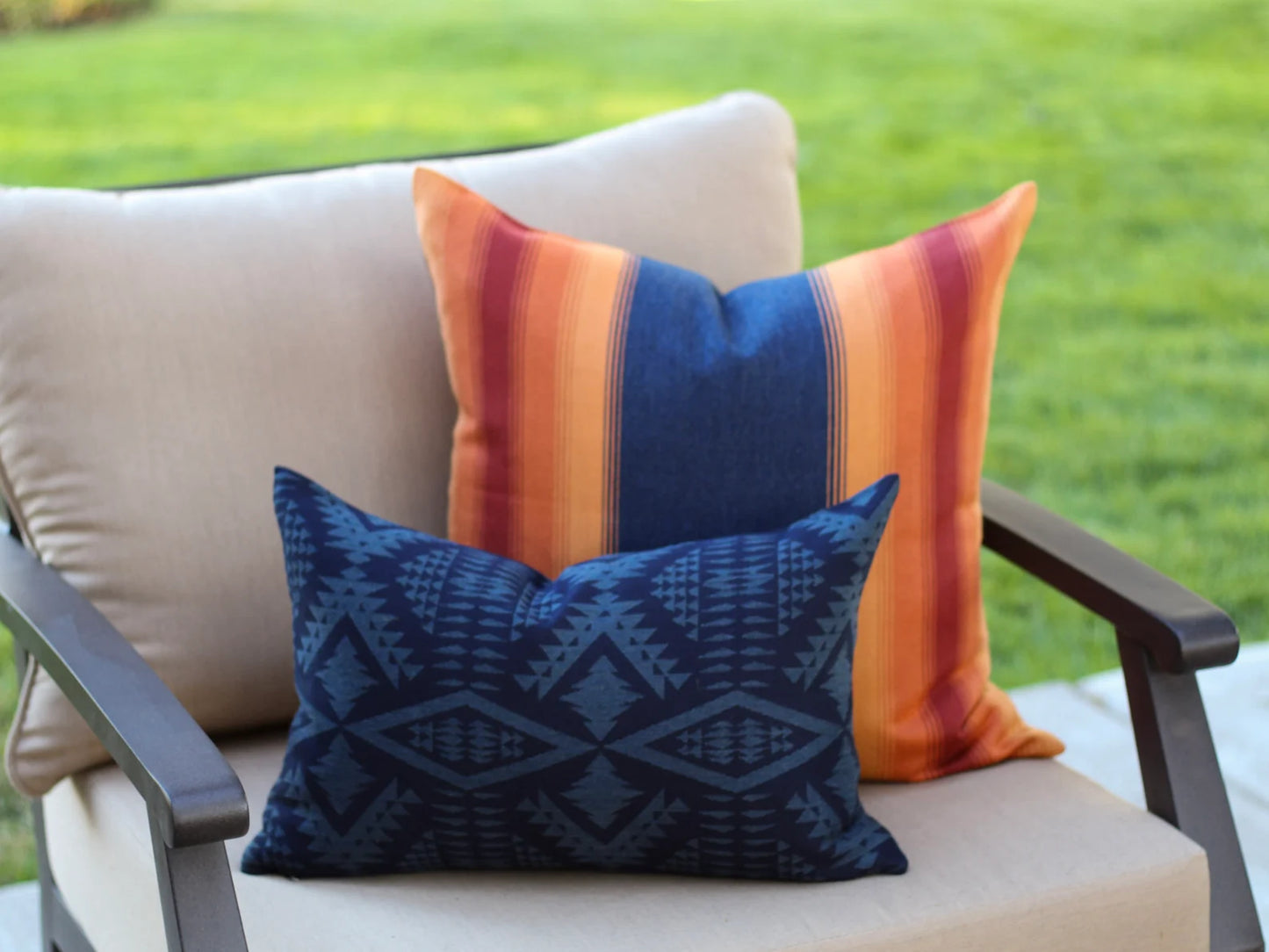 Two decorative outdoor pillows, one in in the two-tone blue River Tonal Midnight pattern and one in the orange, red, and navy Grand Canyon National Park pattern, on a beige outdoor chair.