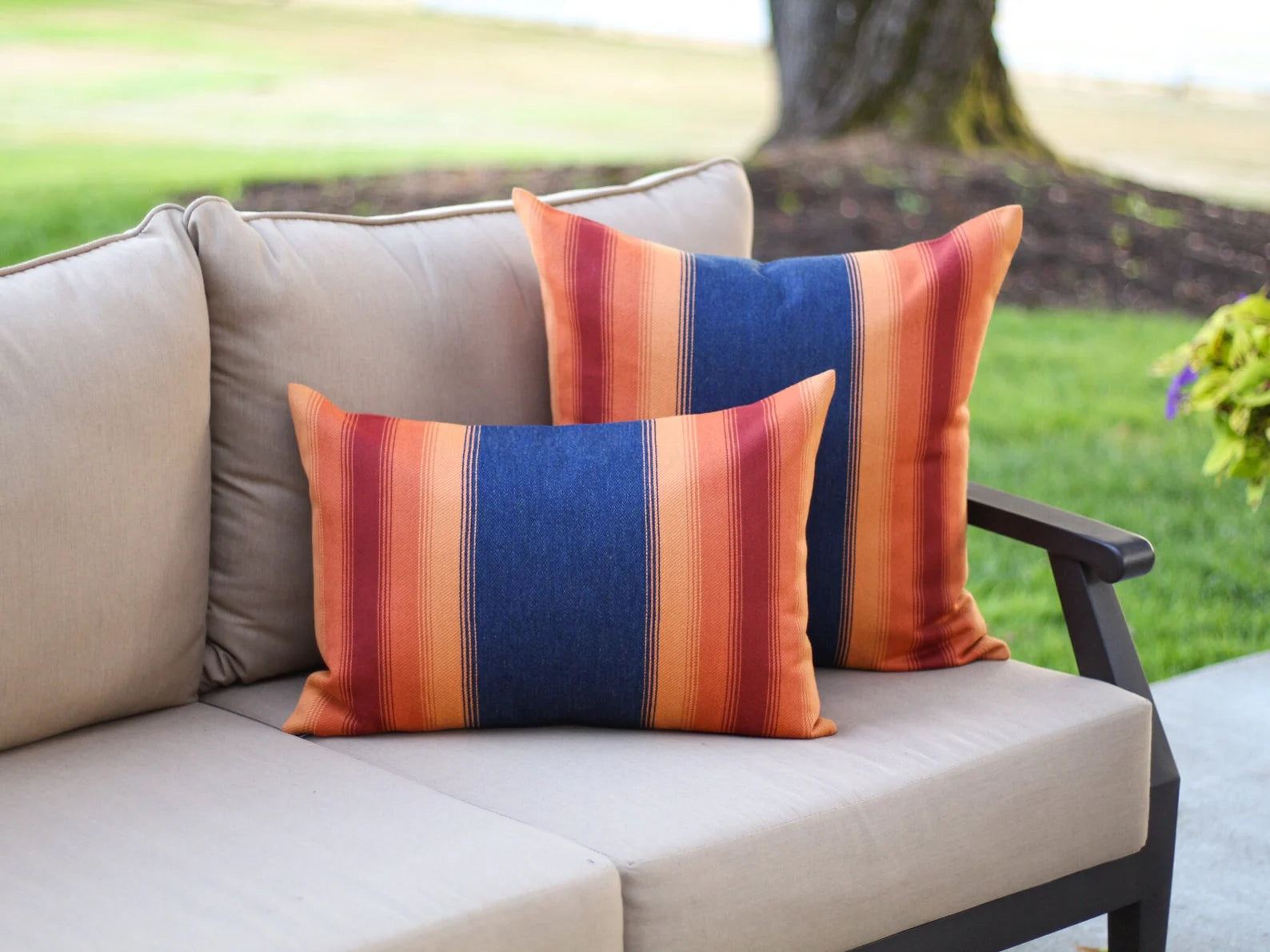 Colorful striped outdoor pillows with a navy background and orange and red stripes on a beige outdoor sofa with a garden background