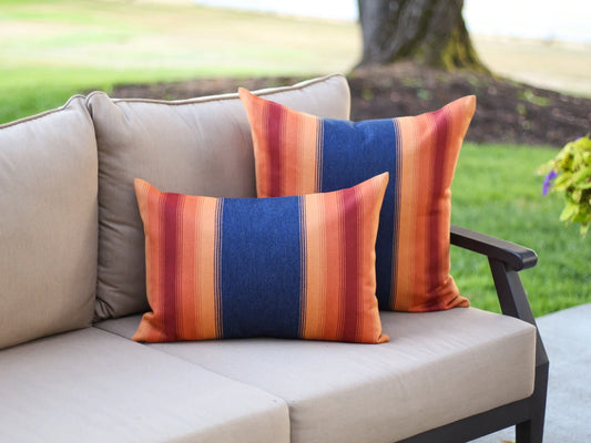 Colorful striped outdoor pillows with a navy background and orange and red stripes on a beige outdoor sofa with a garden background