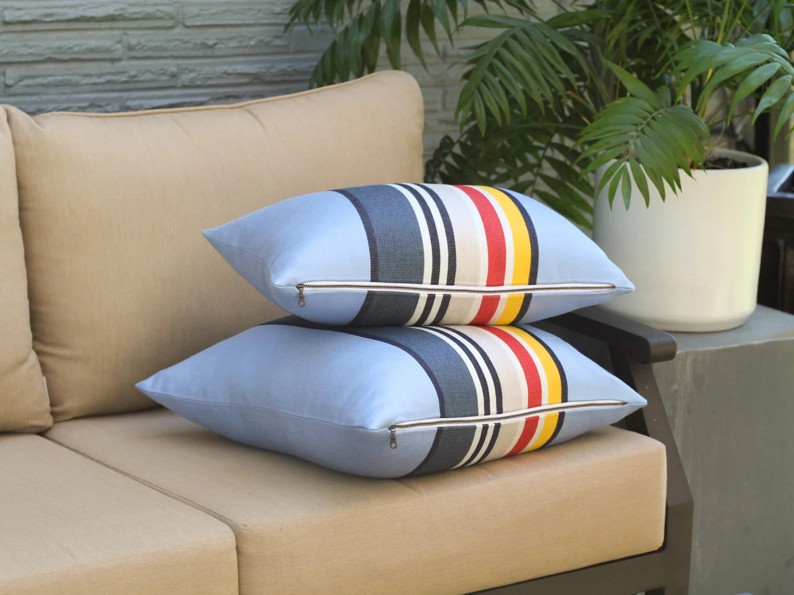 Beige outdoor sofa with outdoor pillow covers in the Yosemite National Park pattern with blue, red, white, and yellow stripes, and a plant in the background