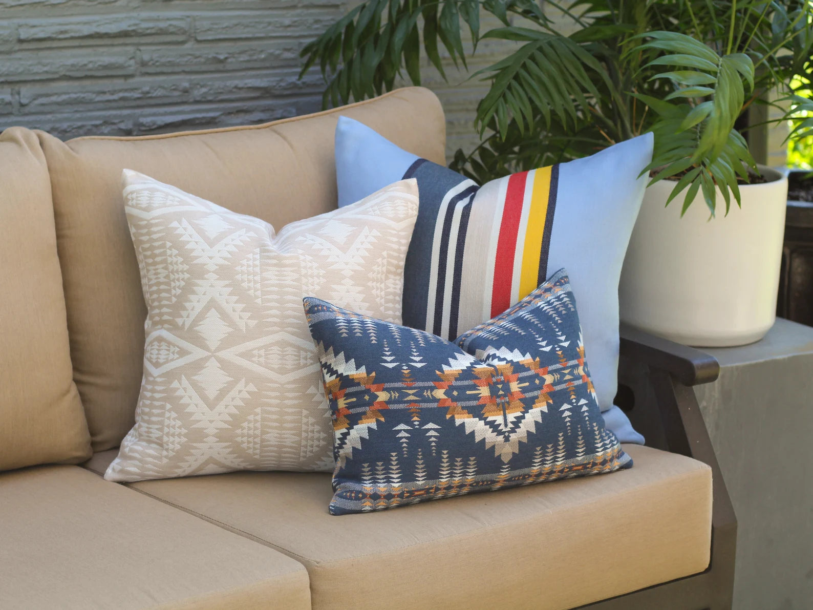 Beige outdoor sofa with patterned outdoor pillows against a stone wall and potted plant.
