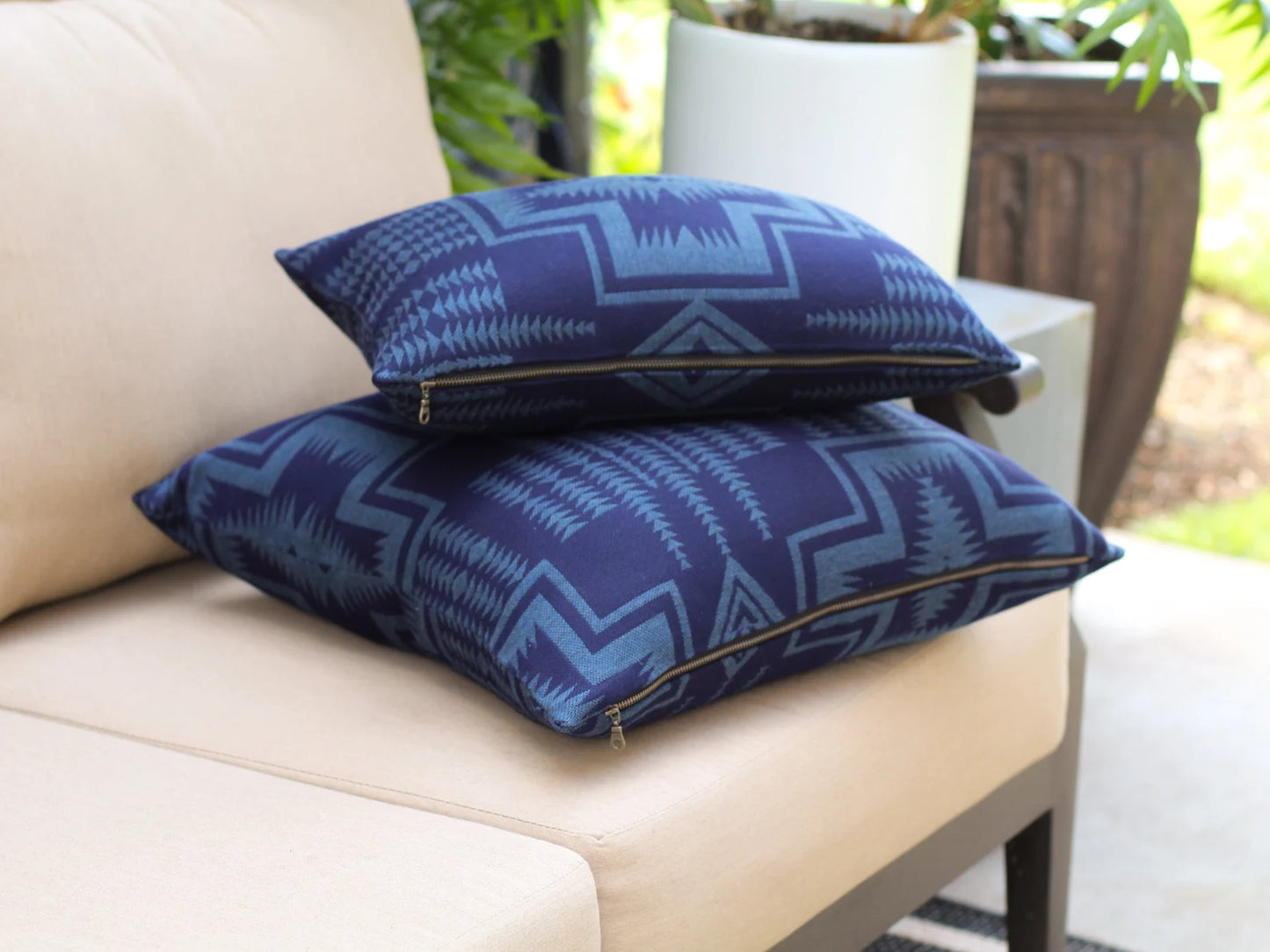 Two outdoor pillows in the two-tone blue Harding Indigo pattern on a beige outdoor sofa with a blurred green background