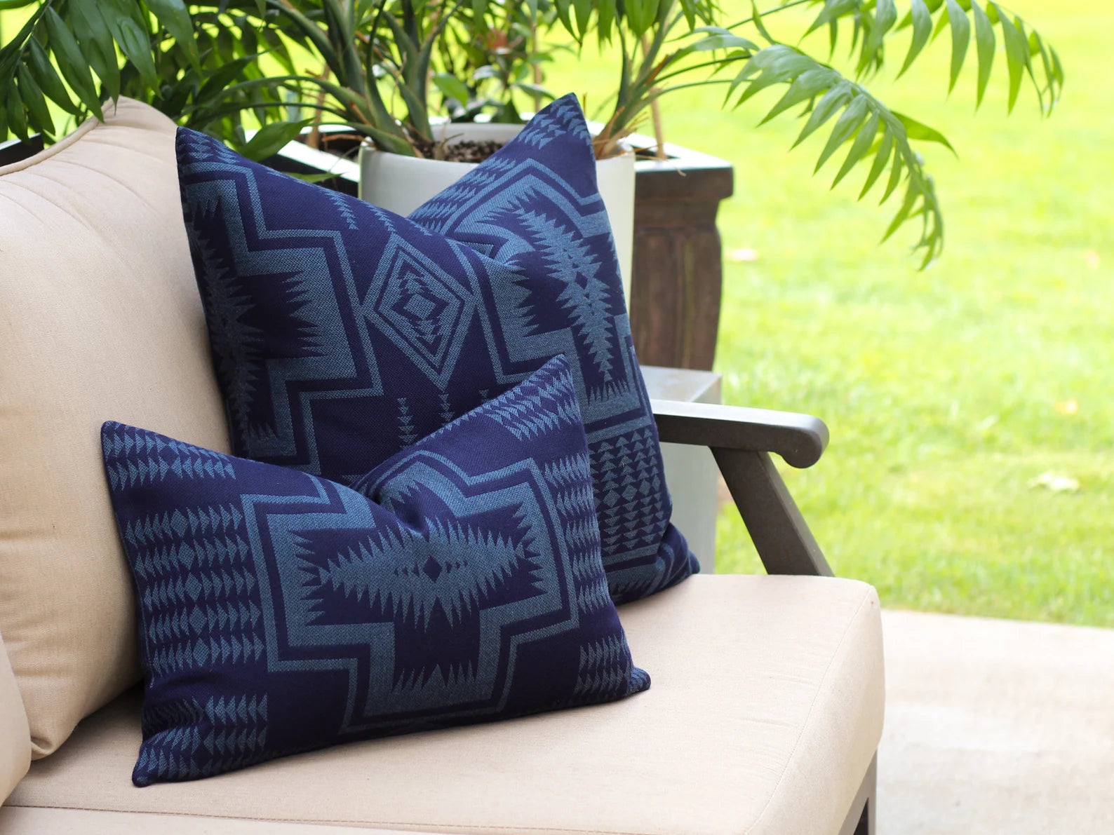 Two outdoor pillows in the two-tone blue Harding Indigo pattern on a beige outdoor sofa with greenery in the background
