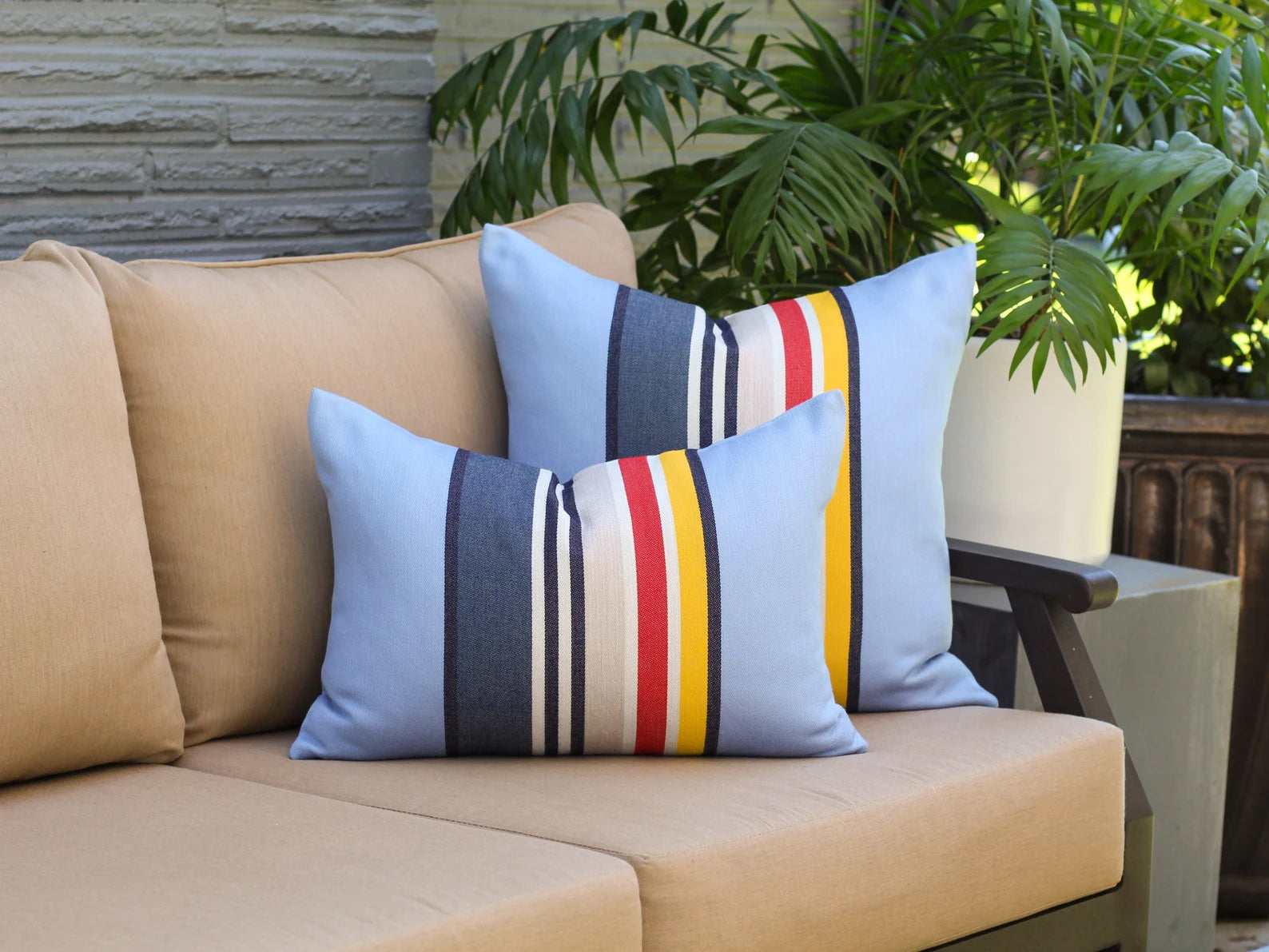 Beige outdoor sofa with striped blue Yosemite National Park outdoor pillows featuring red, yellow, and white stripes, against a stone wall and green plants background.