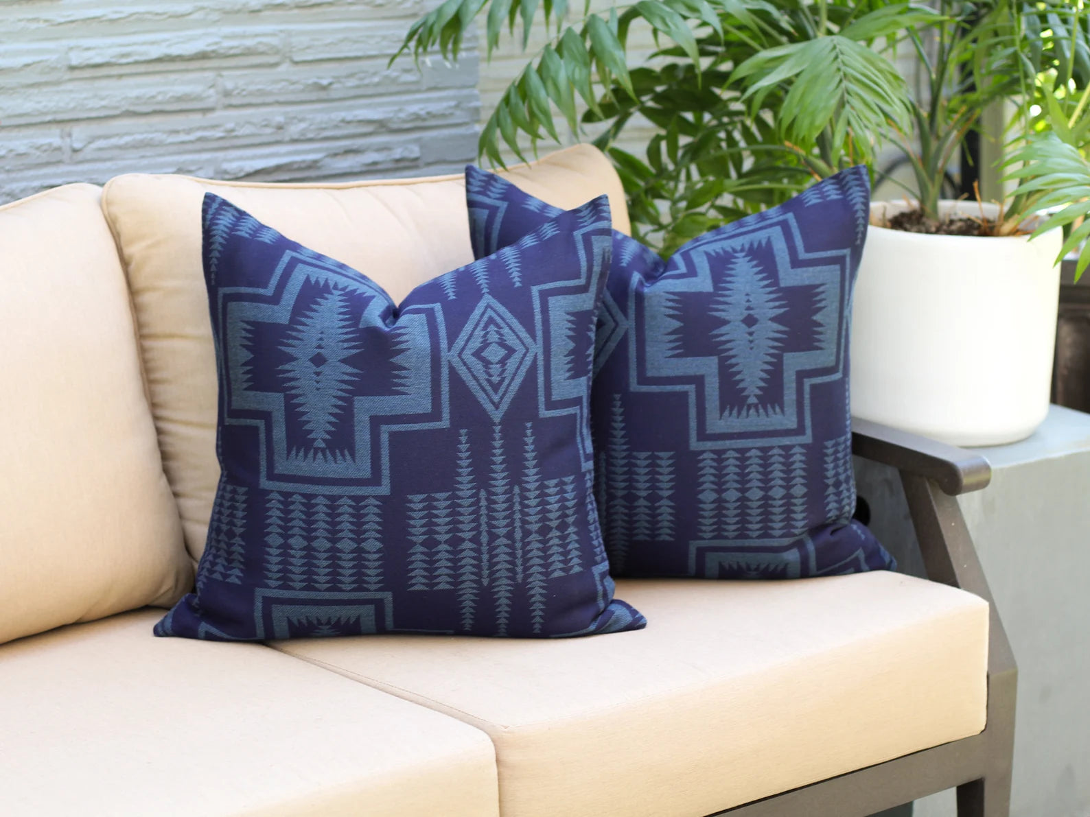 Two outdoor pillows the two-tone blue Harding Indigo pattern on a beige outdoor sofa with plants in the background.