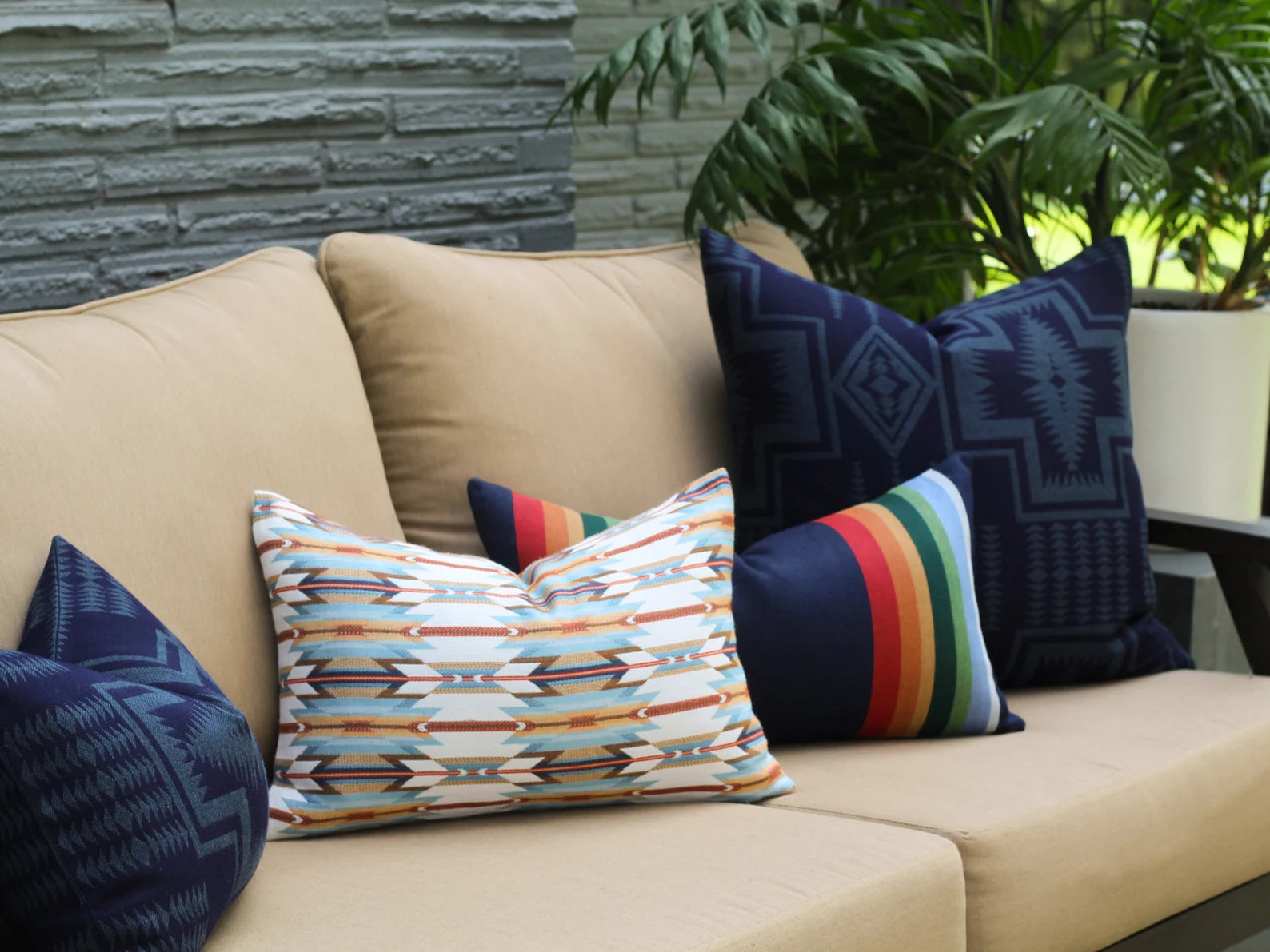 Beige outdoor sofa with patterned outdoor pillows against a stone wall and plants.