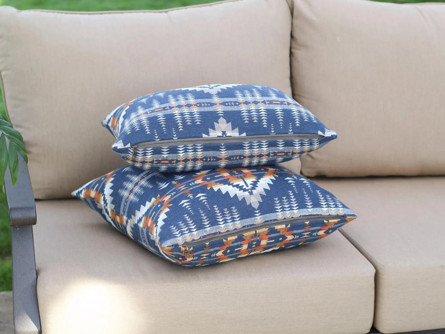 Two outdoor pillows in the Juniper Mesa pattern on a beige outdoor sofa. The Juniper Mesa pattern has a dark blue background with shades of brown, orange, yellow, and white in a southwestern pattern.