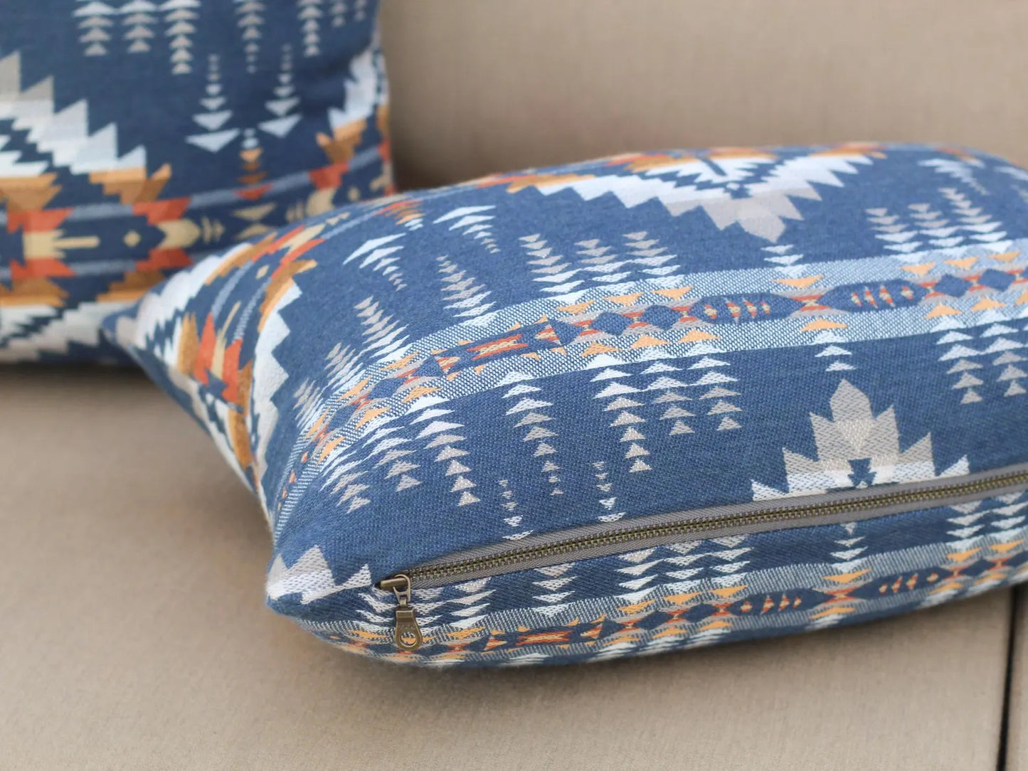 Close up of exposed metal zipper on outdoor pillow in the Juniper Mesa pattern on a beige outdoor sofa. The Juniper Mesa pattern has a dark blue background with shades of brown, orange, yellow, and white in a southwestern pattern.