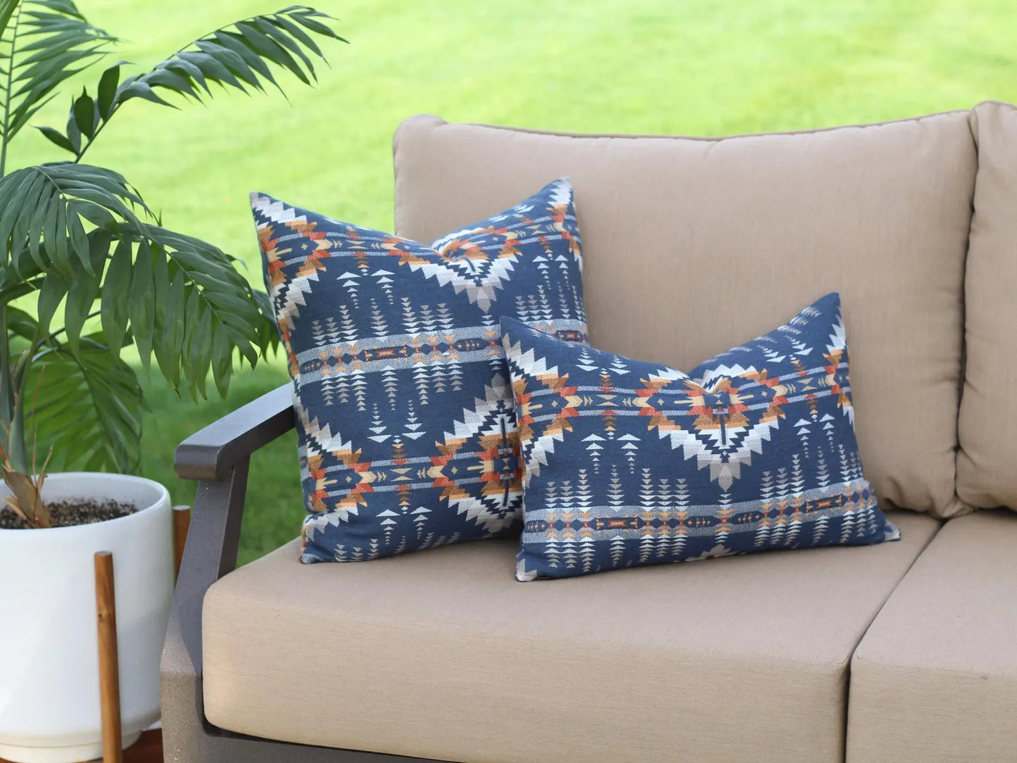 Two outdoor pillows in the Juniper Mesa pattern on a beige outdoor sofa. The Juniper Mesa pattern has a dark blue background with shades of brown, orange, yellow, and white in a southwestern pattern.