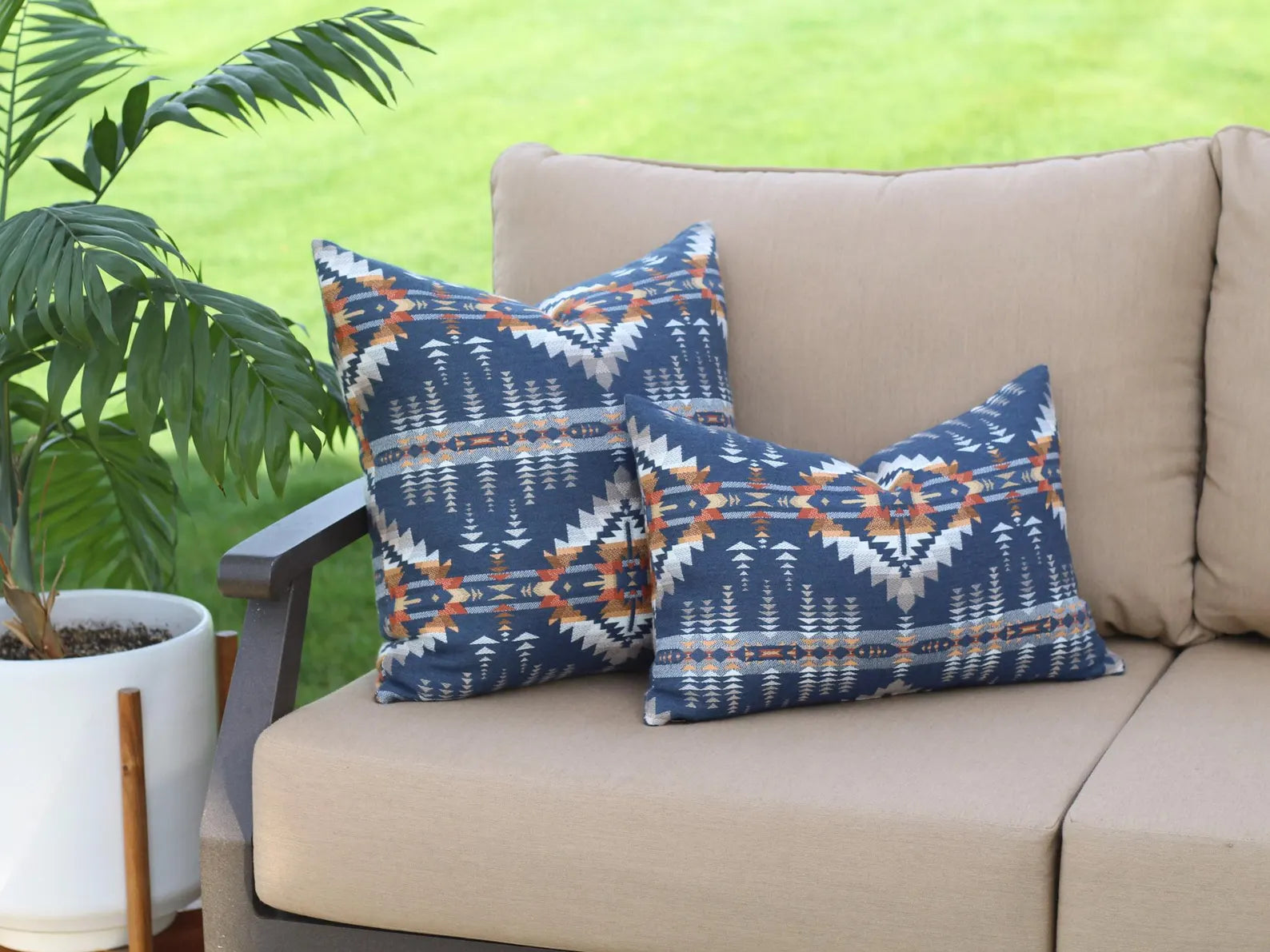 Two outdoor pillows in the Juniper Mesa pattern on a beige outdoor sofa. The Juniper Mesa pattern has a dark blue background with shades of brown, orange, yellow, and white in a southwestern pattern.