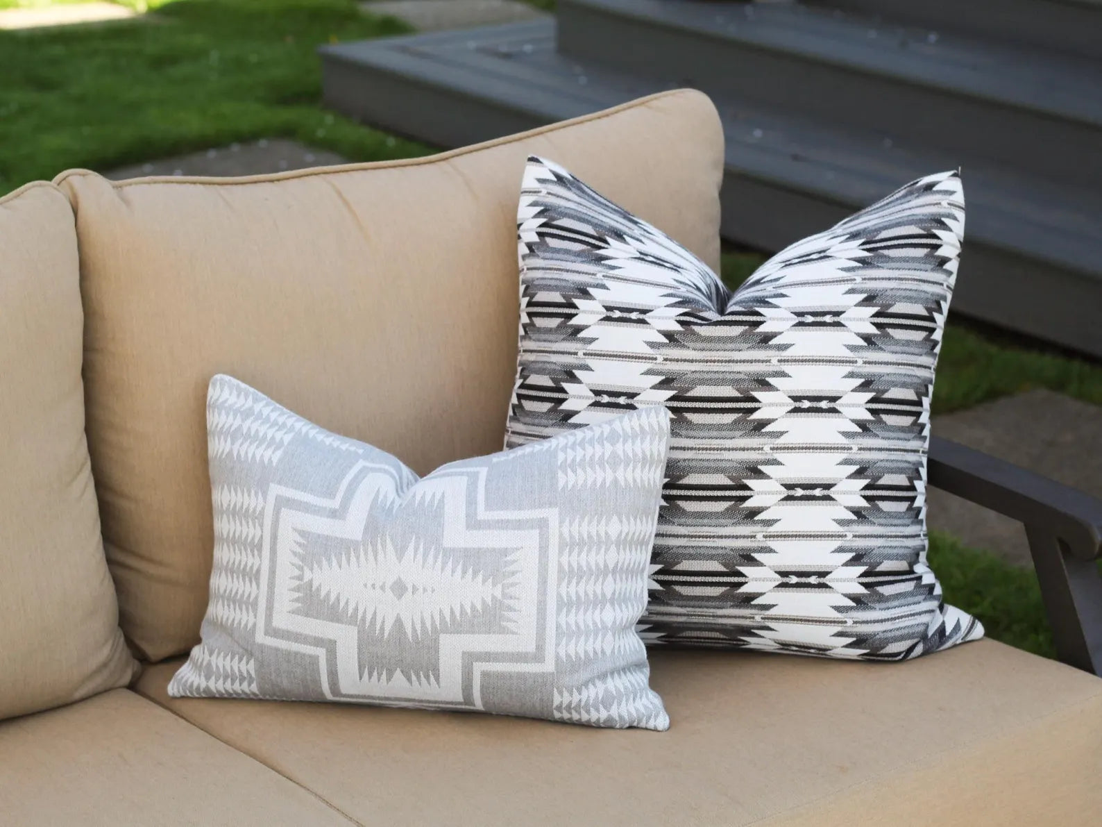 Two outdoor pillows in the Wyeth Trail Stone and Harding Dove patterns on a beige outdoor sofa with a grassy area in the background.