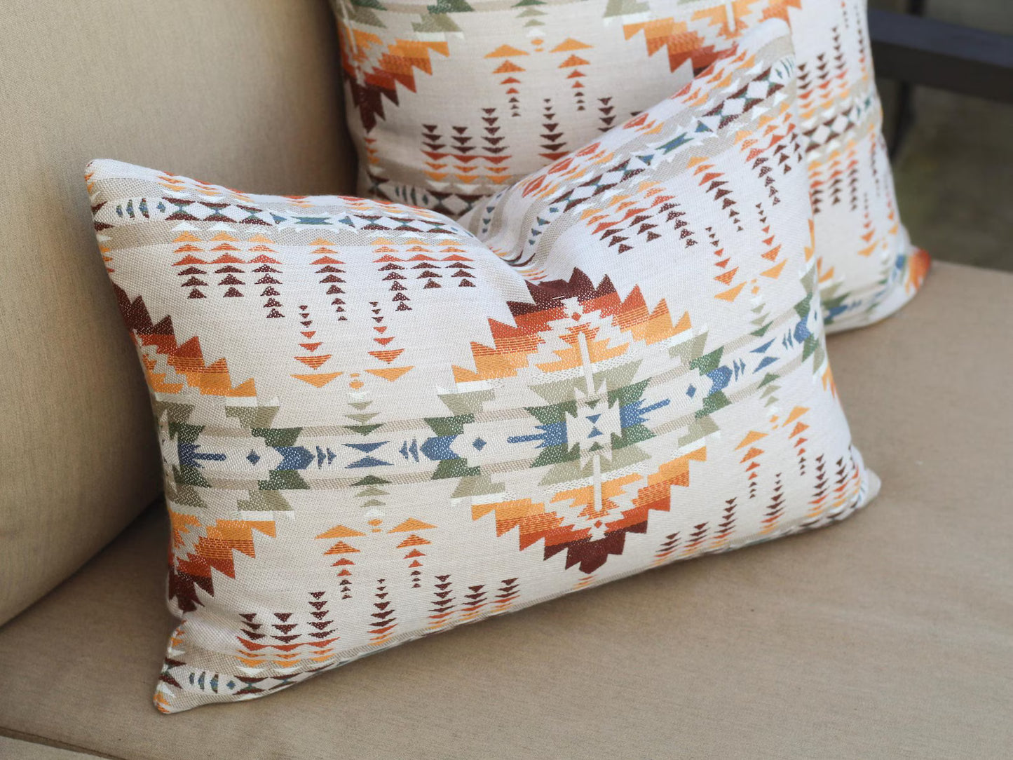 Close up of outdoor pillow covers in the Juniper Mesa Adobe pattern on a beige outdoor sofa. Juniper Mesa Adobe as a beige background with a southwestern pattern in shades of brown, red, orange, yellow, green, and blue.