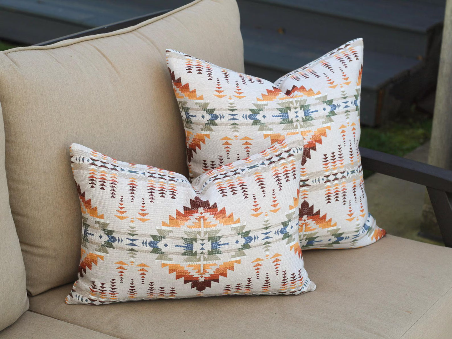 Decorative outdoor pillow covers in the Juniper Mesa Adobe pattern on a beige couch outdoors. Juniper Mesa Adobe as a beige background with a southwestern pattern in shades of brown, red, orange, yellow, green, and blue.