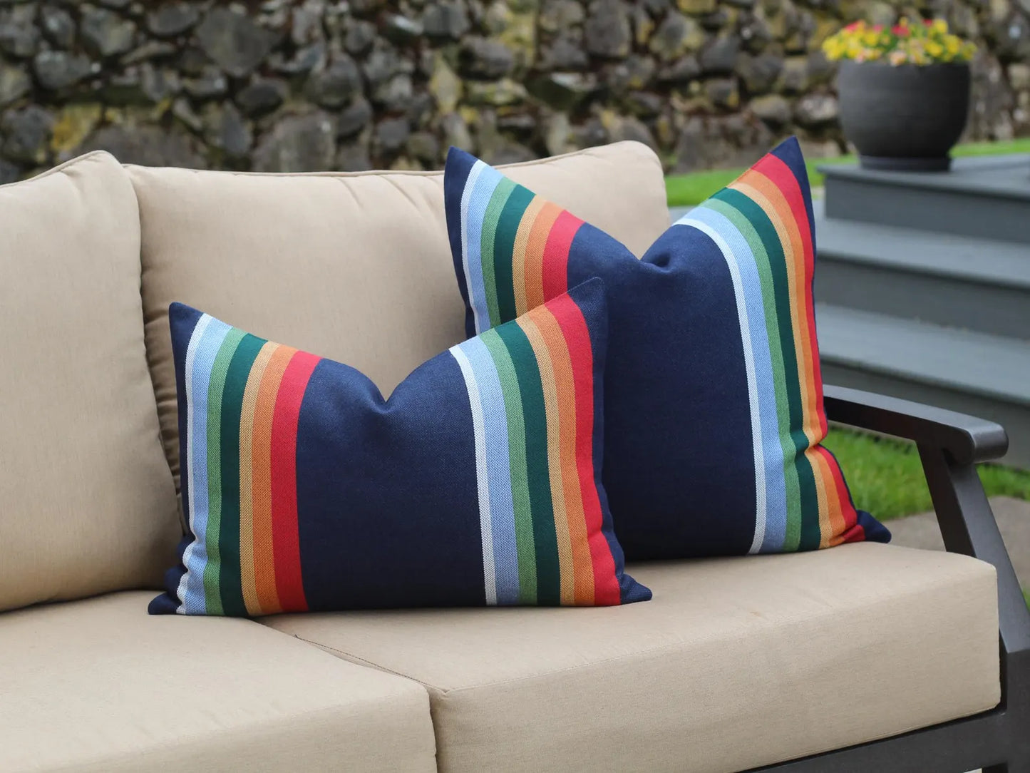 Colorful striped pillows on a beige outdoor sofa with a stone wall and plants in the background.